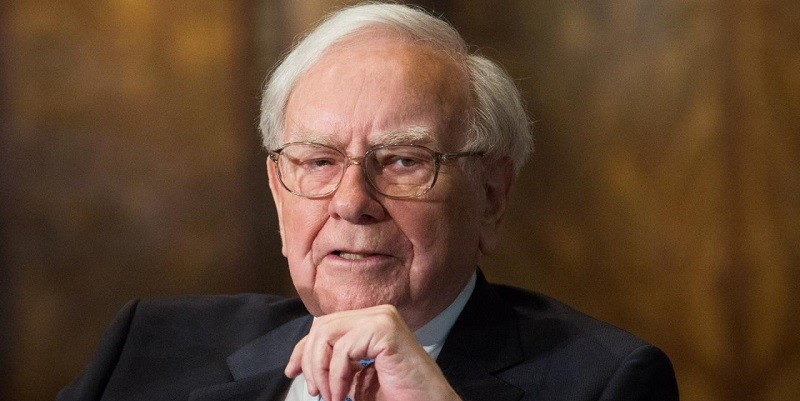 Warren Buffett in pak kijkt serieus. Hij houdt een hand bij zijn kin. De achtergrond is vaag bruin, geen tekst zichtbaar.